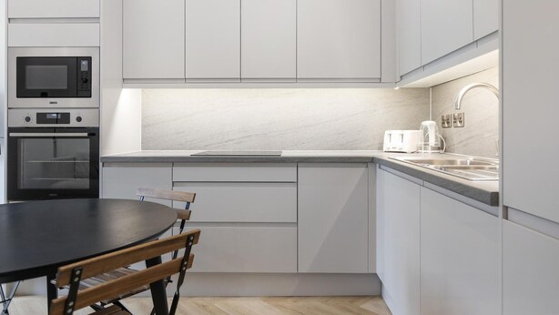 A modern kitchen with white cabinetry, a built-in oven, a microwave, a sink, and a dining table with chairs. - Modern Brixton Flat 3 (Magherafelt)