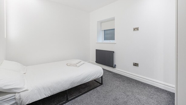 A minimalist bedroom with a single bed, a window, a radiator, and a power outlet. - Modern Brixton Flat 4 (Magherafelt)