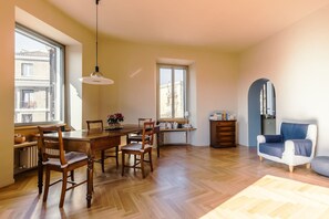 Ein geräumiges Zimmer mit einem Holzespeisetisch, Stühlen, einem blauen Sessel, einem Heizkörper und großen Fenstern.
