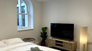 A modern hotel room with a large bed, a flat-screen TV, a small side table, a potted plant, and a floor lamp. - Charming Flat on High Street Ken (London)