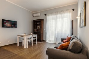 A living room with a sofa, a dining table with chairs, a TV mounted on the wall, and a window with curtains. - 2-bedroom Roman Gem in Rome (Rome)