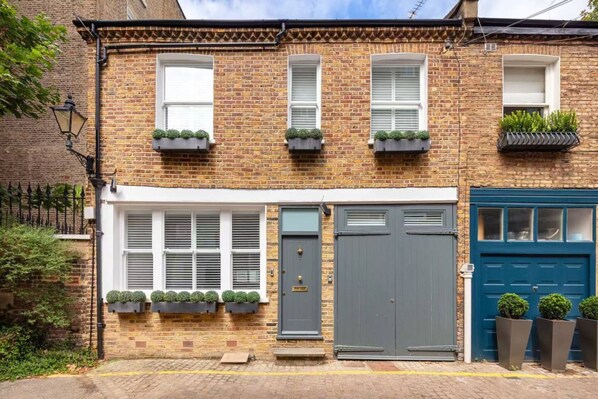 A two-story brick townhouse with a grey front door, white-framed windows, and a blue garage door. - Charming Mews House in Kensington (London)