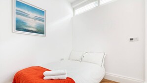 A single-bed room with white walls, a red blanket, and a painting of a beach scene. - Charming Mews House in Kensington (London)