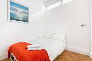 A single-bed room with white walls, a red blanket, and a painting of a beach scene. - Charming Mews House in Kensington (London)