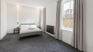 A modern bedroom with a large bed, a radiator, a window with curtains, and a view of buildings outside. - Spacious Flat in Trendy Brixton (London)
