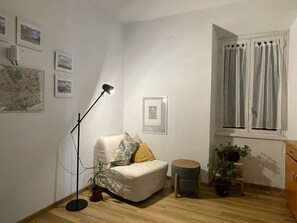 Ein Zimmer mit einer Couch, einer Stehlampe, einer Karte, einem Fenster mit Vorhängen und einem kleinen Hocker.