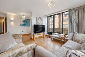Uma sala de estar com televisão, sofás, um banco e janela com vista para prédios.