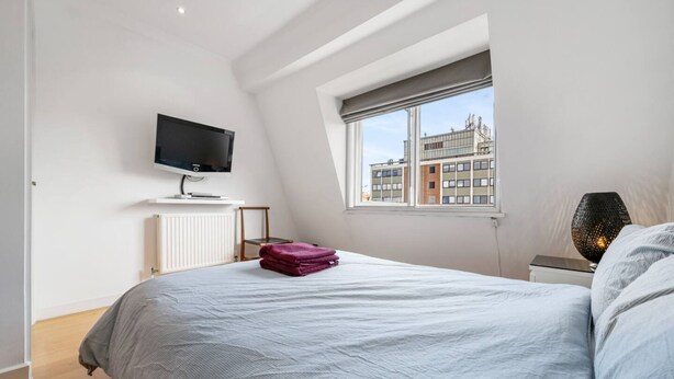 A bedroom with a bed, a TV mounted on the wall, a window with a view of buildings, and a bedside table with a lamp. - Spacious Kensington Apartment With City Views (London)