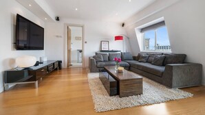 A modern living room with a grey sofa, a coffee table, a flat-screen TV mounted on the wall, and a large window providing a view of the city. - Spacious Kensington Apartment With City Views (London)