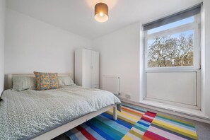 Ein Schlafzimmer mit Bett, Heizkörper, Kleiderschrank und Fenster mit Blick auf Bäume.