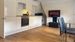 Ett modernt kök med vita skåp, en inbyggd ugn och en matplats med ett svart bord och stolar. En stor tv-skärm som visar "NETFLIX" är monterad på väggen.