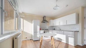 A modern kitchen with white cabinets, a round table with wooden legs, a built-in stove, and a window with blinds. - 4-bed Maisonette With Balcony (London)