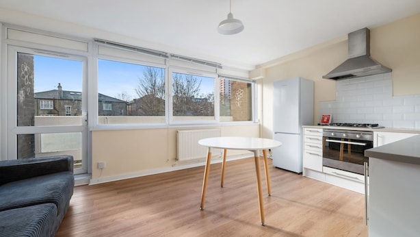 A modern kitchen with a white stove, oven, and cabinets, a round wooden table, and large windows offering a view of the outside. - 4-bed Maisonette With Balcony (London)