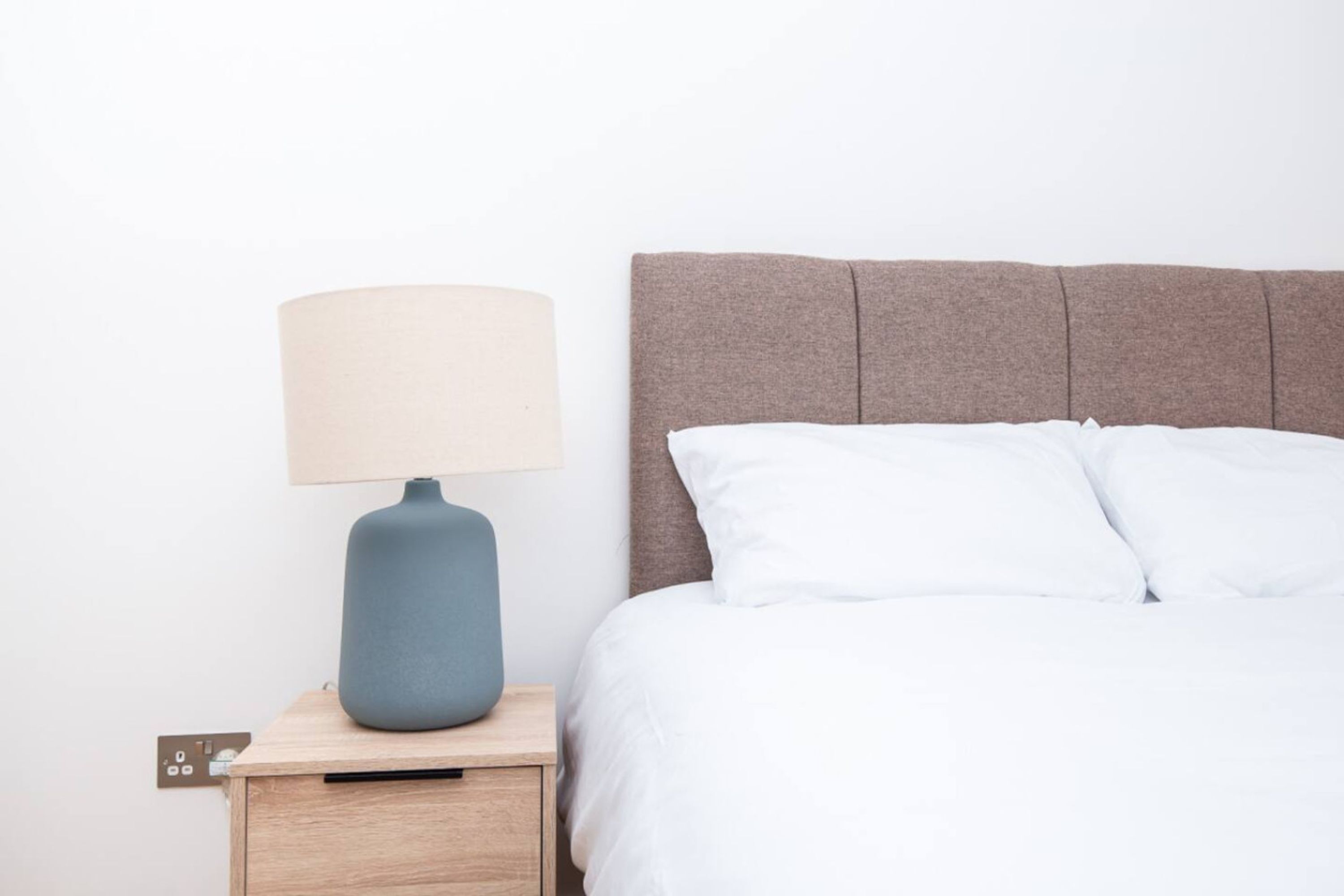 Un lit avec une literie blanche, une tête de lit grise et une table de chevet avec une lampe.