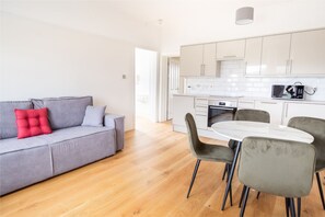 A modern living room with a grey sofa, a red cushion, and a dining area with a white table and grey chairs. - 2BR Seaside Sanctuary in St Leonards on Sea Flat 5 (St Leonards-On-Sea)
