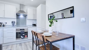 A modern kitchen with white cabinets, a stainless steel range hood, and a dining area with a wooden table and chairs. - Penny Place Retreat Central 2bed 2bath by Station Free Parking (Ashford)