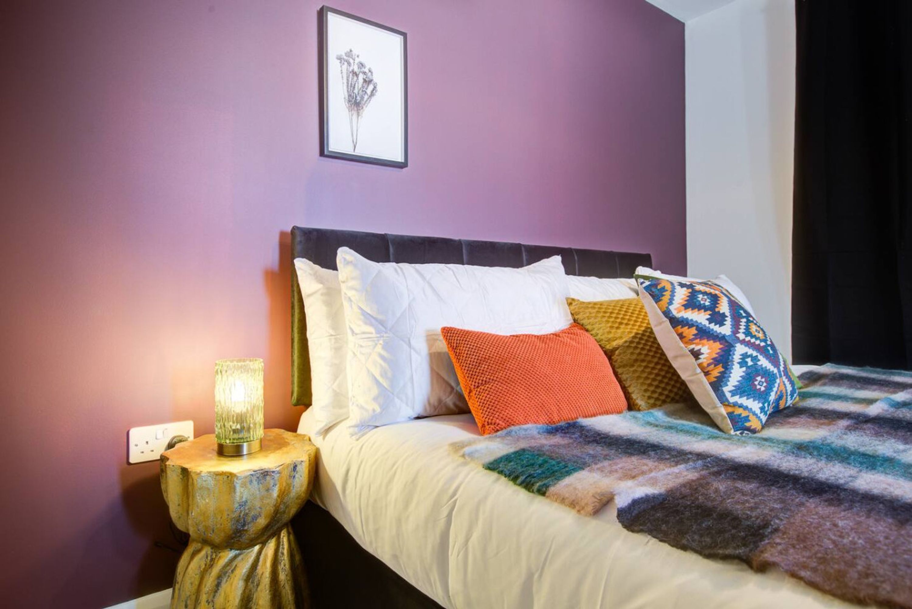 Una cama con almohadas blancas y estampadas, una manta multicolora y una mesita con una lámpara.