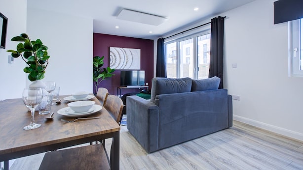 A modern living room with a grey sofa, a dining table with plates and glasses, and a TV in the background. - Penny Place Retreat Central 2bed 2bath by Station Free Parking (Ashford)