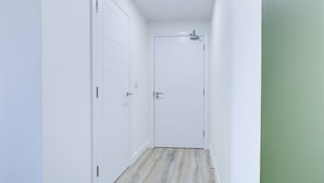 A narrow hallway with white walls and a wooden floor, leading to a white door with a small window. - Penny Place Retreat Central 2bed 2bath by Station Free Parking (Ashford)