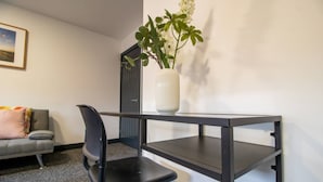 Una habitación con un escritorio de metal negro, una silla de oficina negra, un jarrón blanco con hojas verdes, un sofá gris con un cojín rosa y un cuadro en la pared.