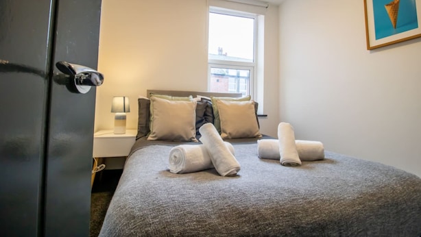 Una cama bien tendida con almohadas y toallas enrolladas, una mesita de noche con una lámpara y una ventana con vistas a edificios.