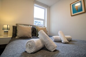 A neatly made bed with rolled white towels, a bedside lamp, and a window with a view of a building outside. - Cozy 1BR Flat Nestled in the Heart of Sunderland (Sunderland)
