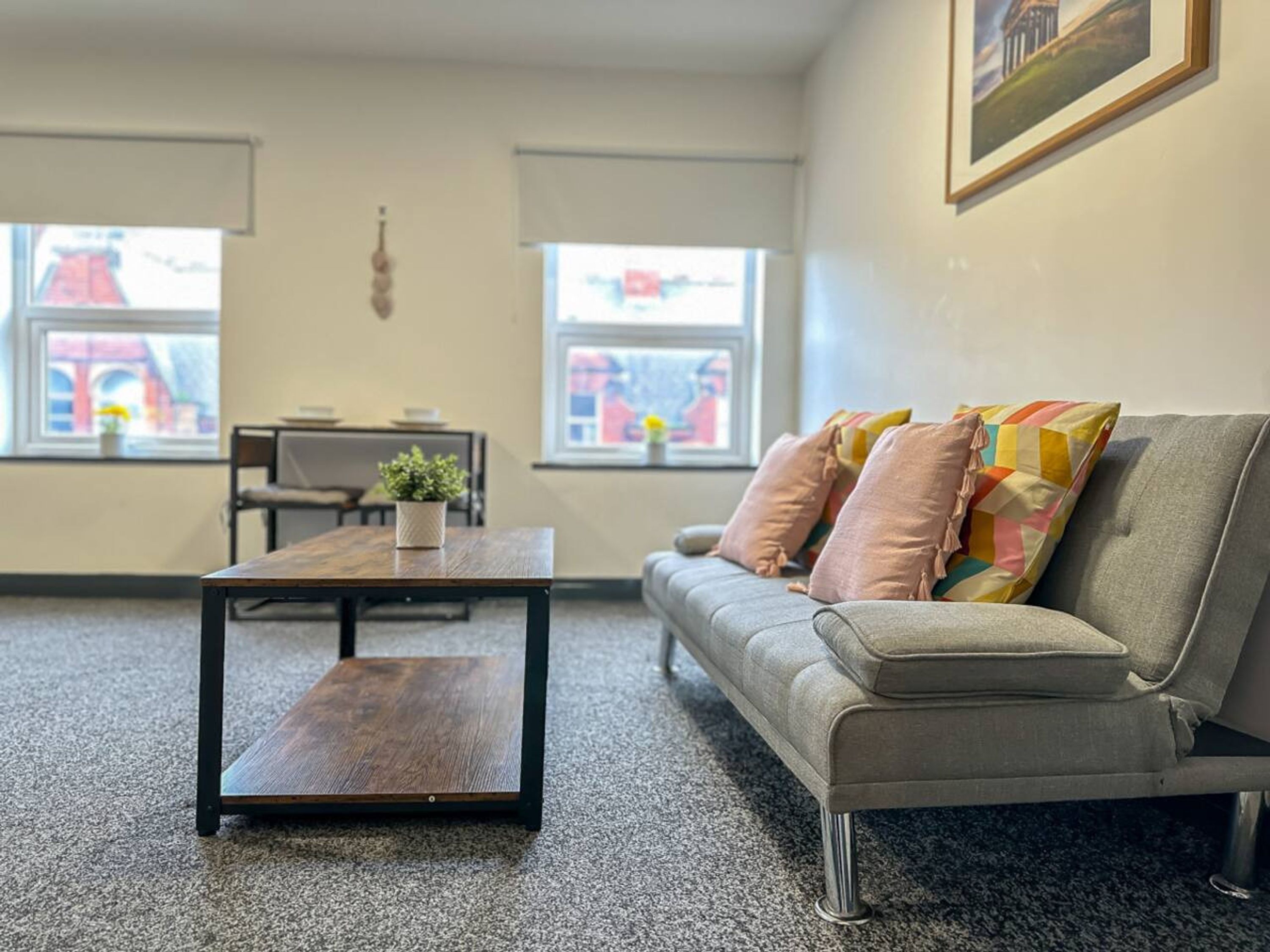 A modern living room with a sofa, a coffee table, and a small plant. There are two windows with a view of a building outside.