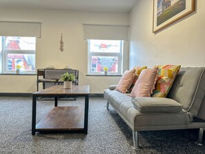 Uma sala de estar moderna com um sofá, uma mesa de centro e uma pequena planta. Há duas janelas com vista para um edifício.