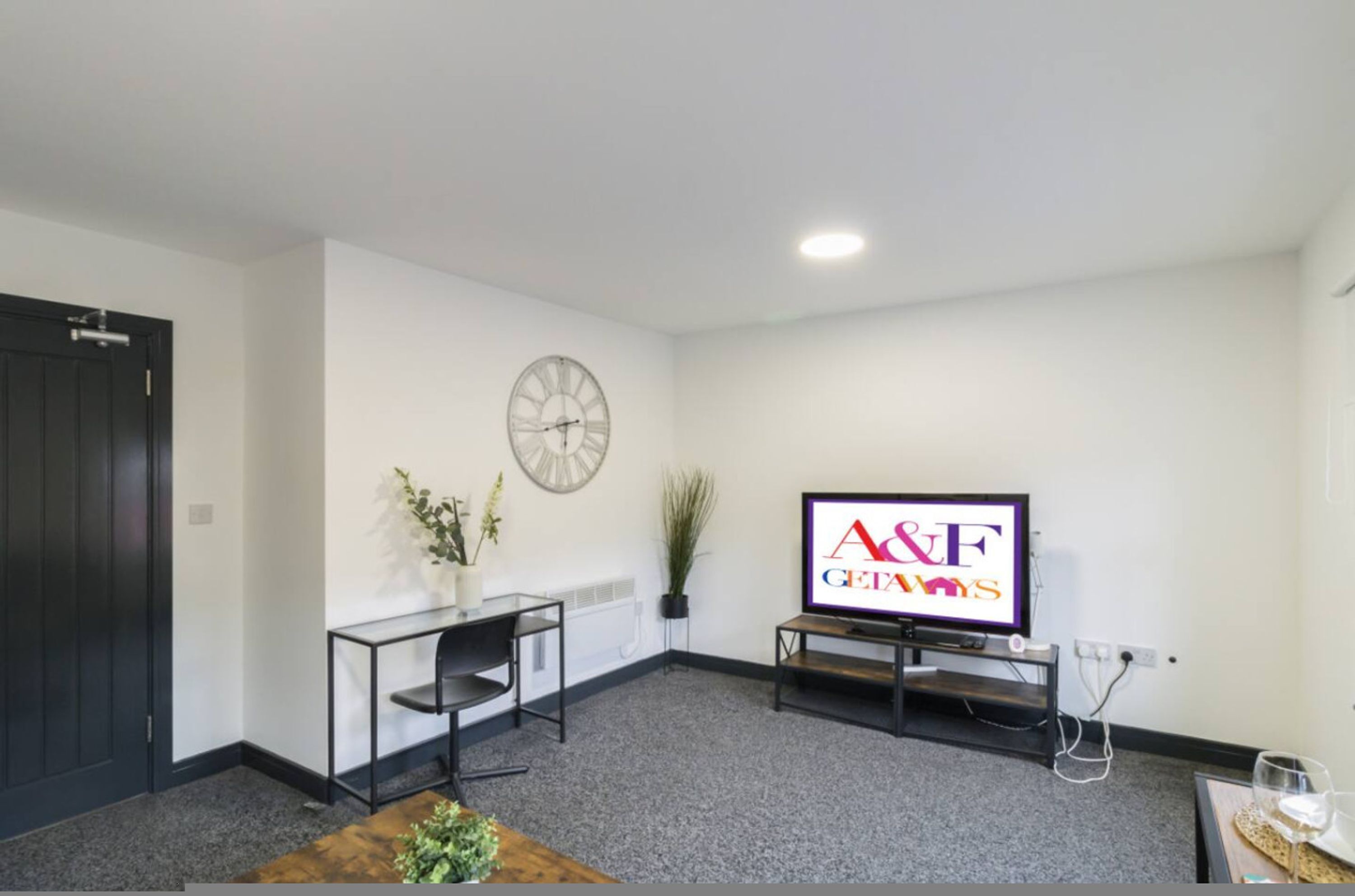 Een kamer met een televisie waarop 'A&F GETAWAYS' staat, een klok, een kleine tafel met een stoel, en een plant.