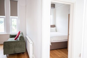 Una habitación con un sofá verde, una cama con ropa de cama blanca y una ventana con persianas.