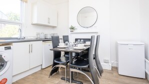 A kitchen with white cabinets, a dining table set with chairs, a washing machine, and a small refrigerator. - Gloucester Street Nest - 3BR Flat for 7 Guests (Stroud)