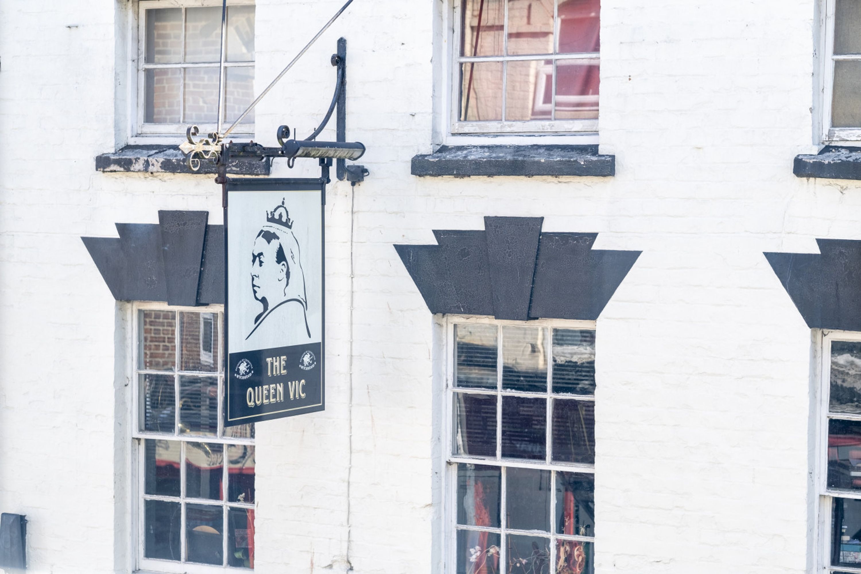 En bygning med et skilt, der viser 'The Queen Vic', og en sort-hvid illustration af en kvinde.