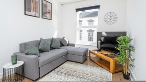A modern living room with a grey sectional sofa, a wooden coffee table, a TV, a potted plant, and a patterned rug. - Gloucester Street Nest - 3BR Flat for 7 Guests (Stroud)