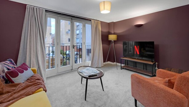 A hotel room with a sofa, armchair, a small table with a magazine, a TV on a stand, and a large window with curtains. - Harland Haven Spacious 4BR House in Ipswich (Ipswich)