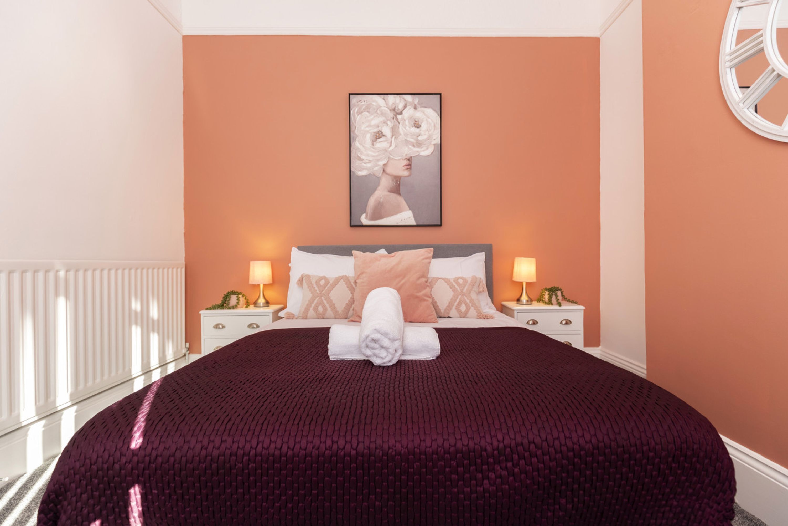 Una cama bien hecha con una toalla enrollada, dos lámparas de noche, una obra de arte enmarcada en la pared y un aparador blanco con cajones.