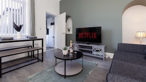 A living room with a television displaying 'NETFLIX,' a sofa, a coffee table with a bottle and glasses, and a dining area with benches. - Newly Renovated 3BR House Near Hospital uni (Sunderland)