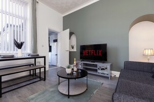 Una sala de estar con una televisión mostrando 'NETFLIX', un sofá, una mesa de centro con una botella y vasos, y un comedor con bancos.