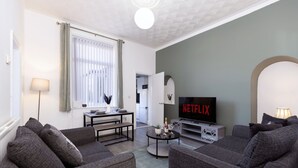A living room with grey sofas, a coffee table, a TV displaying Netflix, a lamp, and a window with blinds. - Newly Renovated 3BR House Near Hospital uni (Sunderland)