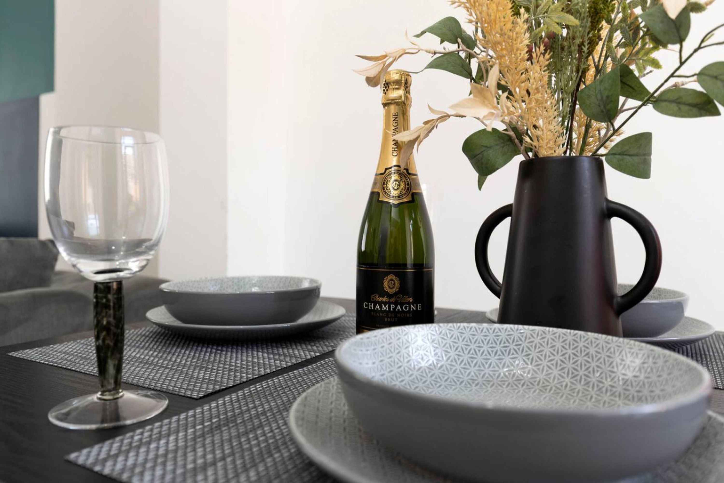 Uma mesa de jantar com uma garrafa de champanhe, uma taça e um vaso com plantas secas.