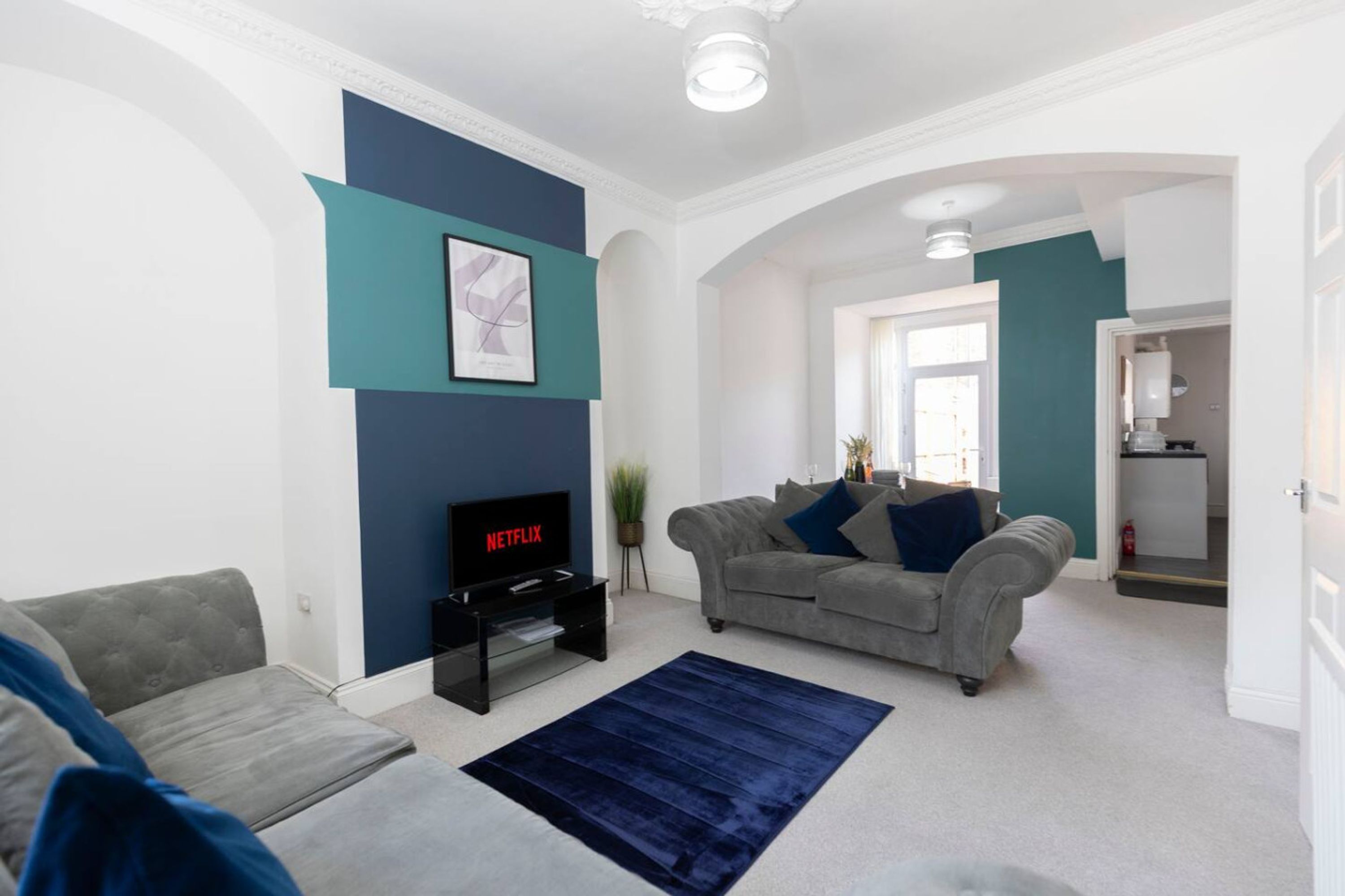 Un salon avec un canapé gris, un tapis bleu, un téléviseur affichant le logo « NETFLIX » et un mur d’accent de couleur turquoise.