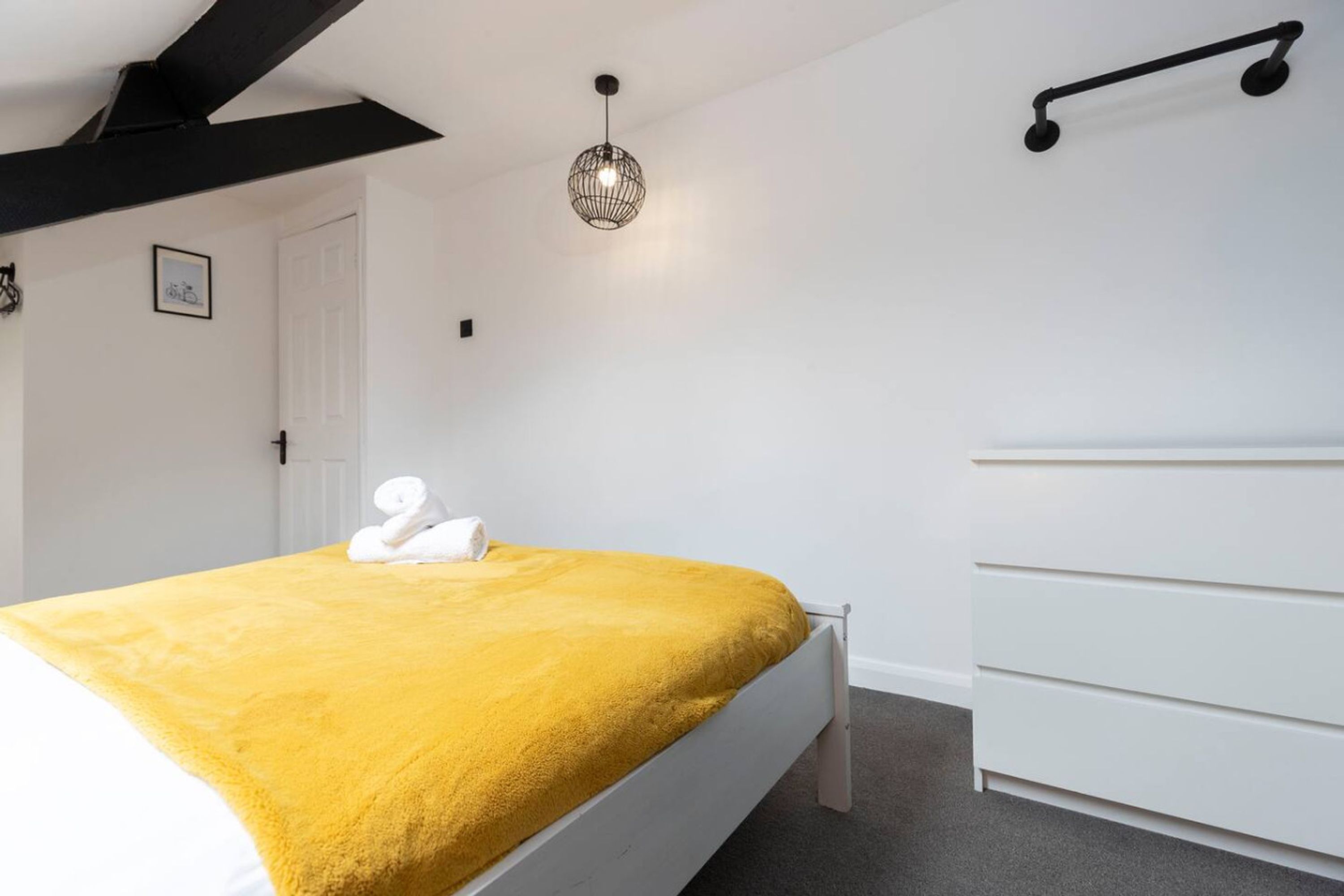 Ein Schlafzimmer mit einem Bett, einer weißen Kommode, einem schwarzen Wandregal und einer Pendelleuchte.