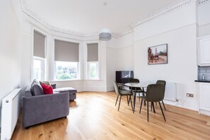 A modern living room with a grey sofa, dining table, and chairs, large windows, and a painting on the wall. - Seaside Sanctuary in St Leonards on Sea - Flat 3 (St Leonards-On-Sea)