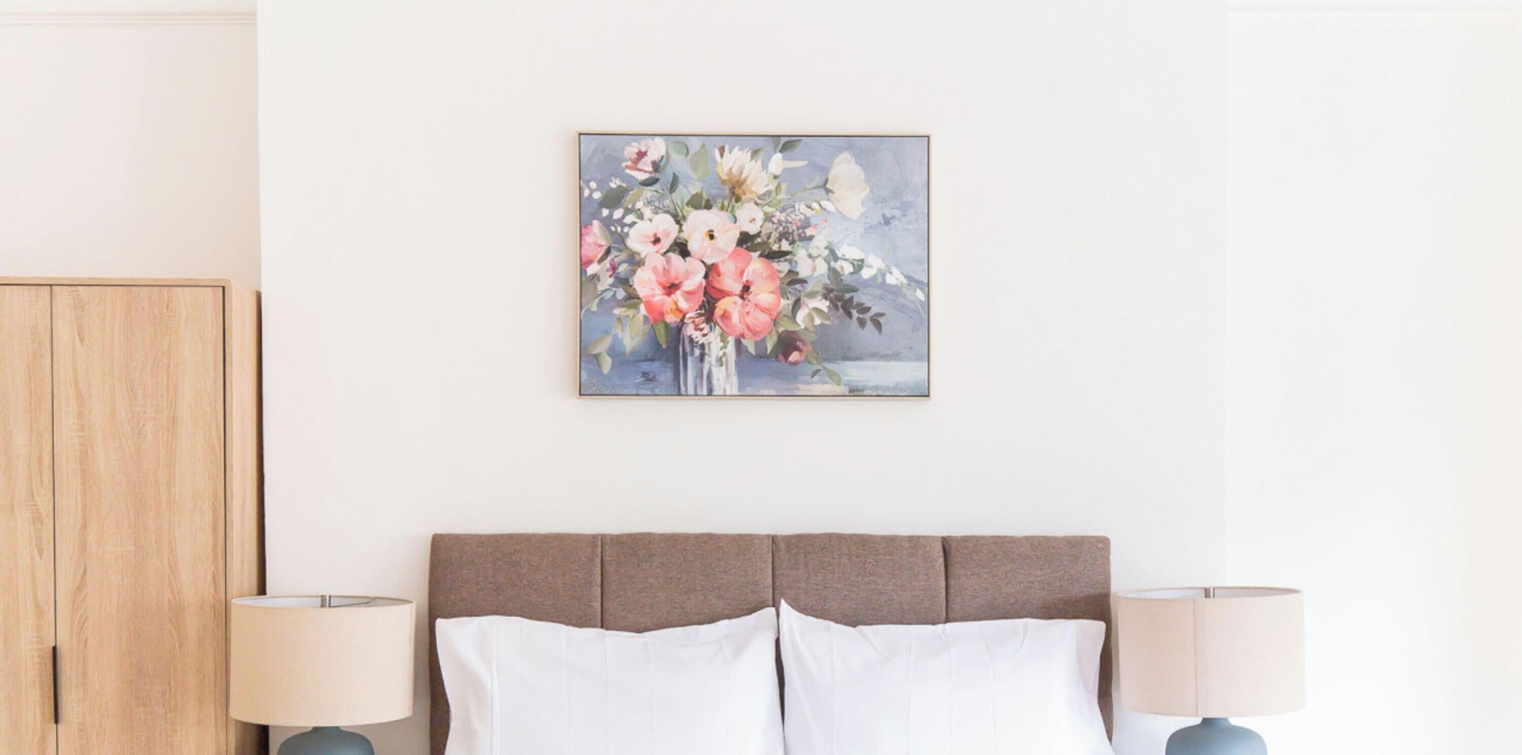 Un dormitorio con una cama, dos lámparas de noche, un armario de madera y un cuadro con motivos florales en la pared.