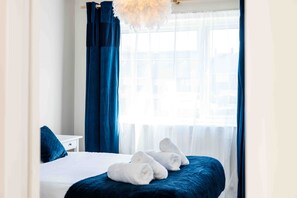 Ein Hotelzimmer mit einem Bett, einem weißen Nachttisch, einer blauen Bettdecke und einem Fenster mit durchsichtigen Vorhängen.