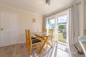 Ein Zimmer mit Esstisch aus Glas, Holzstühlen, einer weißen Tür und Blick auf Grün hinter einem Fenster.