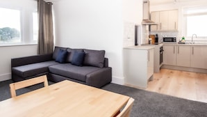Una sala de estar moderna con un sofá gris, una mesa de madera y una cocina con armarios blancos y un microondas.