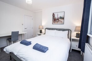 A hotel room with a bed, a desk, a chair, a nightstand, a lamp, and a picture on the wall. - Coastal Charm 1BR Flat Near the Sea Street Parking (St Leonards-On-Sea)