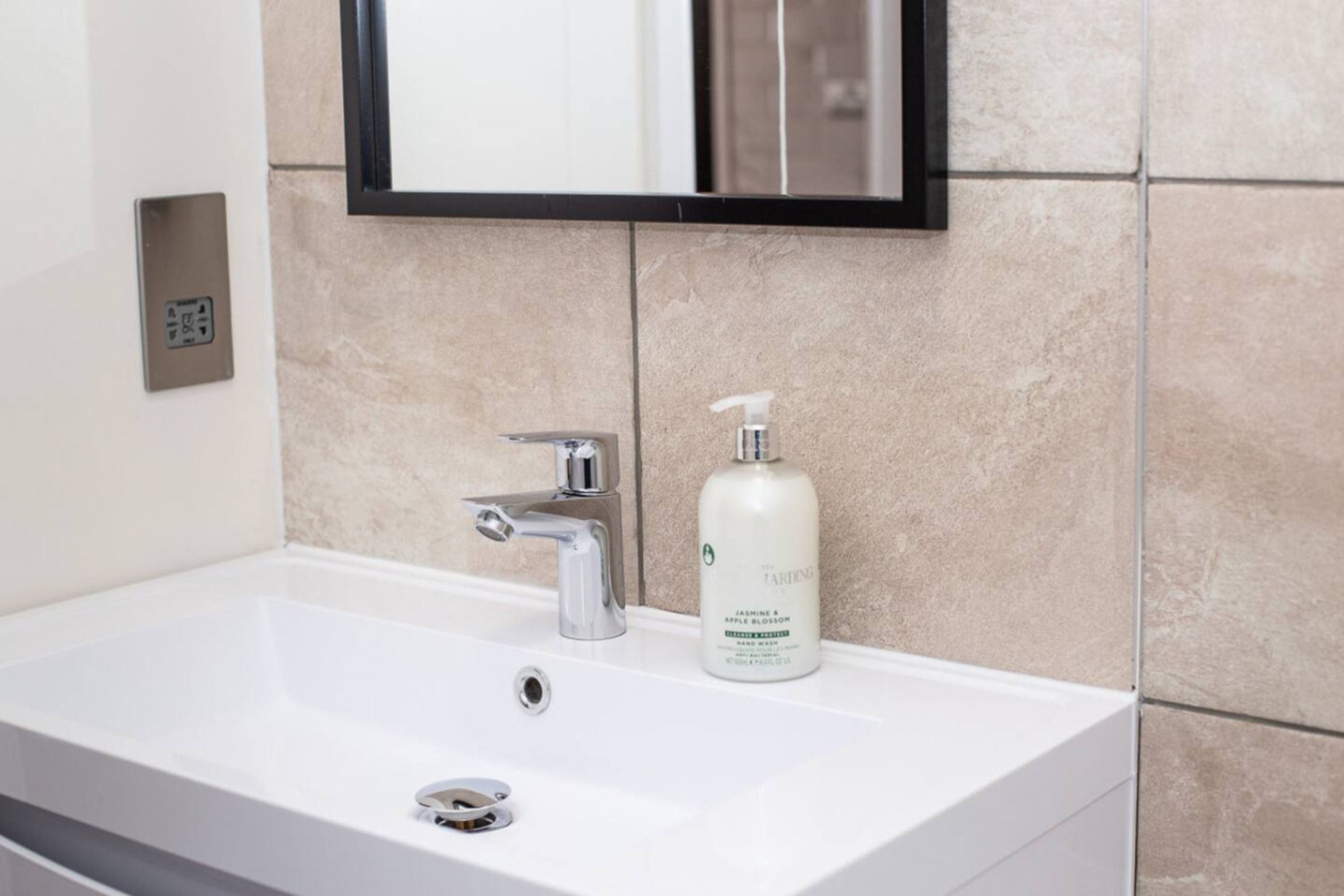 Un baño con un lavamanos blanco, un espejo encima y una botella de loción en el lavamanos.