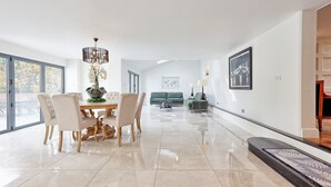 A spacious living room with a dining area, large windows, a chandelier, and a green sofa. - Mayrose House 6BR 4bath Luxe Spacious Retreat (Bishop's Stortford)