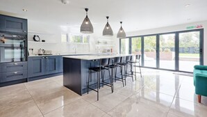 A modern kitchen with dark blue cabinets, a central island with bar stools, pendant lighting, and a large sliding glass door to the outdoors. - Mayrose House 6BR 4bath Luxe Spacious Retreat (Bishop's Stortford)